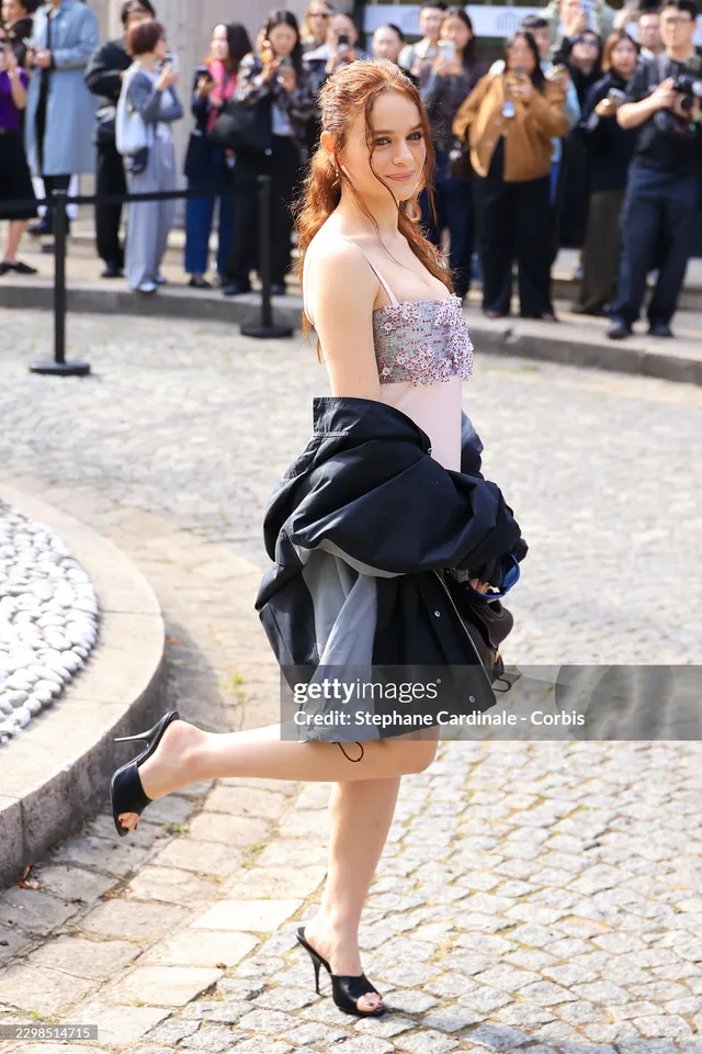 Joey King raising one heel while posing in a pale dress outdoors.