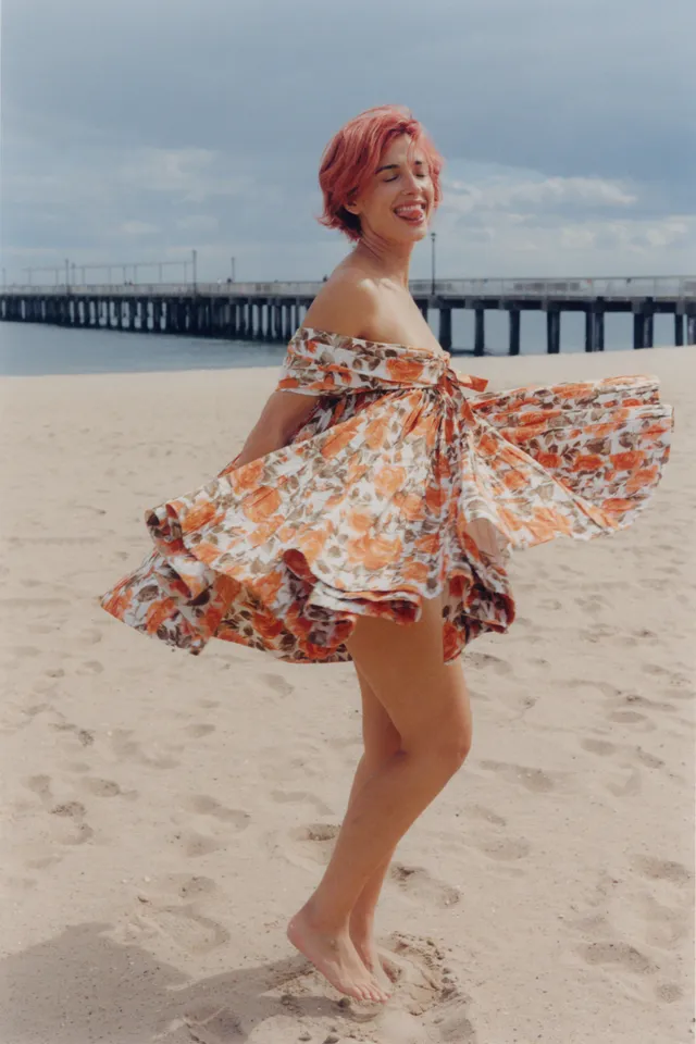 Naomi Scott standing barefoot on a beach in a patterned dress.
