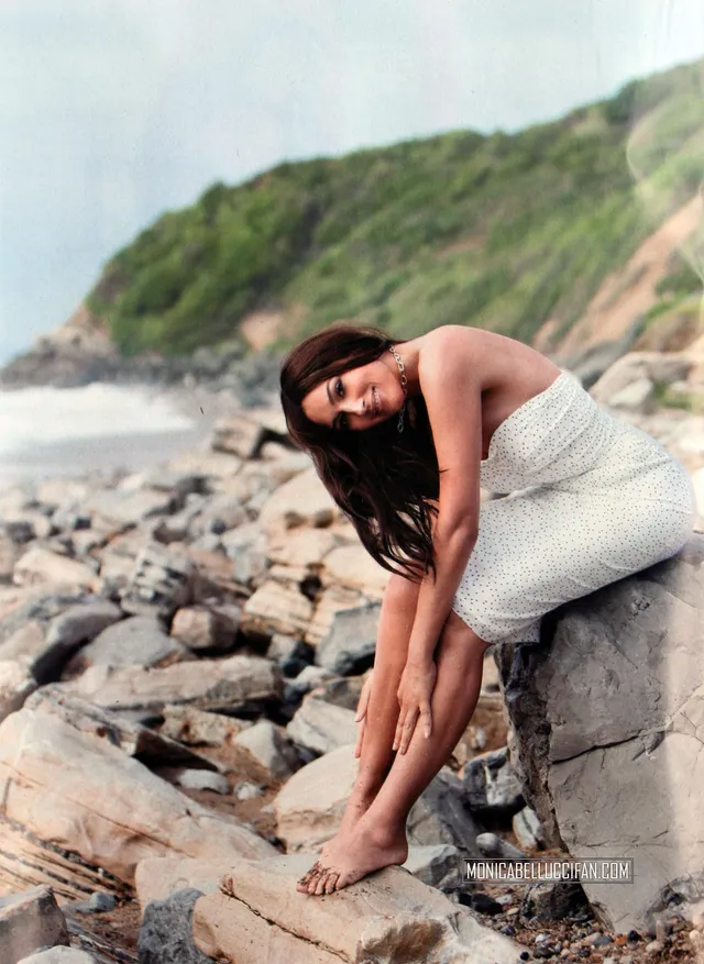 Monica Bellucci seated on seaside rocks in a pale dress.