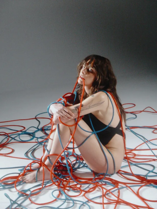Cara Delevingne sitting among colourful cables in a studio photograph.