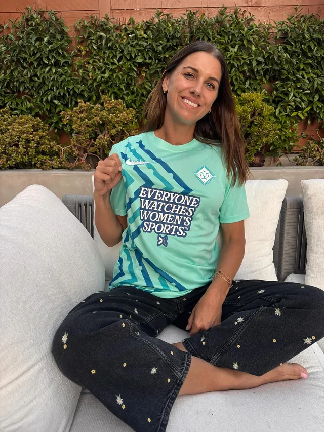 Alex Morgan sitting cross-legged and barefoot on a sofa in a green T-shirt.