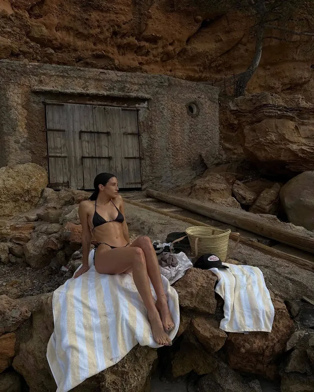 María Pedraza sitting barefoot on a towel beside a rock wall in a bikini.