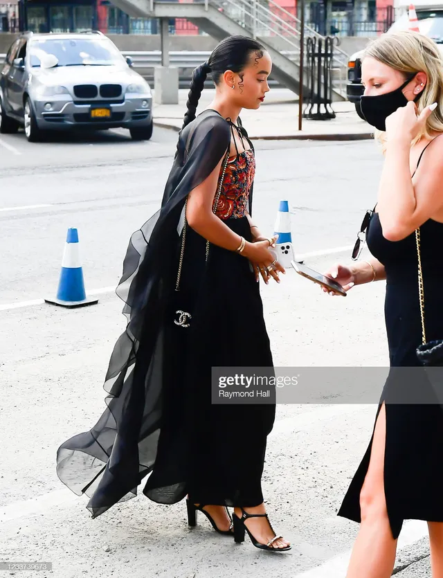 Whitney Peak standing outdoors in a dark skirt, sheer jacket and heeled sandals.