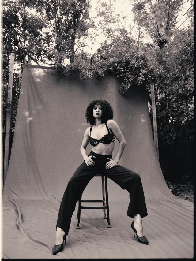 Samantha Logan sitting on a stool in a black outfit and pointed heels.