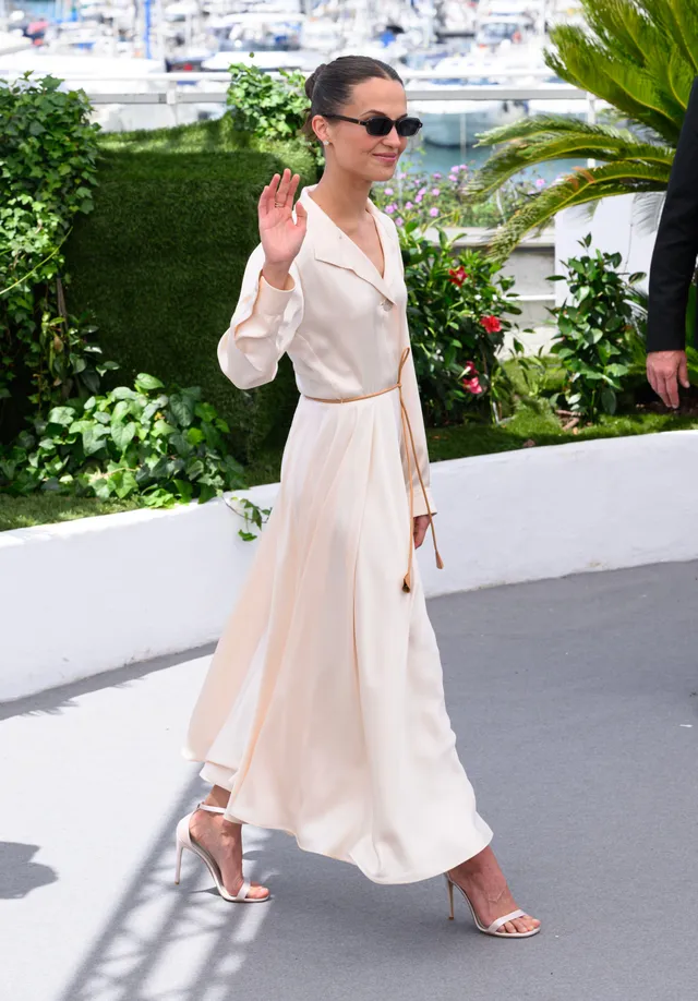 Alicia Vikander waving outdoors in a pale outfit.