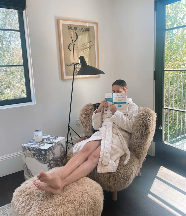 Ashley Tisdale sitting barefoot in a soft chair while reading.