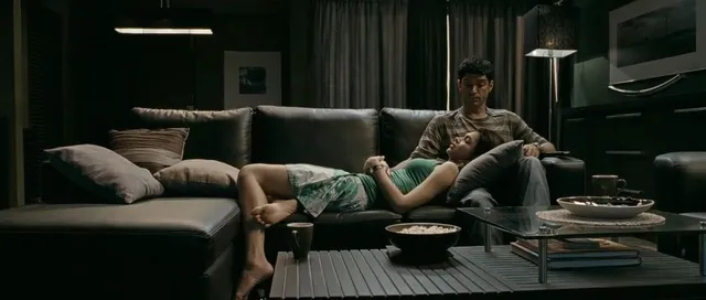 Deepika Padukone reclining barefoot on a sofa in a dimly lit screen scene.