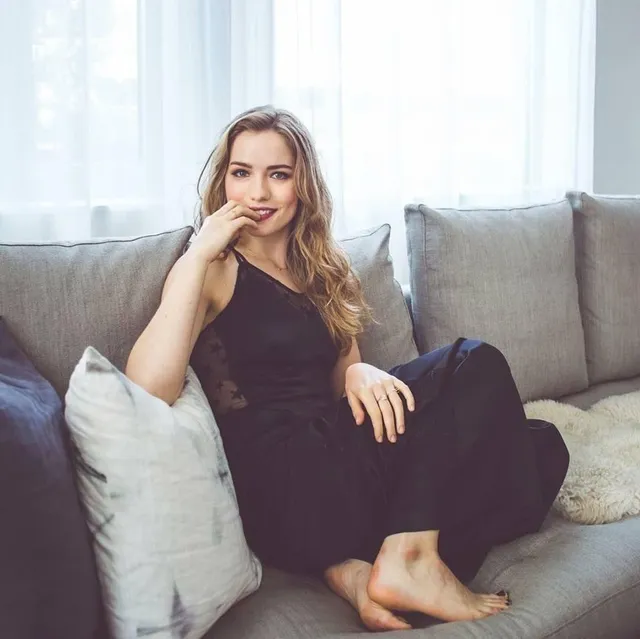 Willa Fitzgerald sitting barefoot on a sofa in a dark dress.