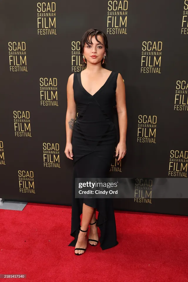 Isabela Merced posing on a red carpet in a black dress and heels.