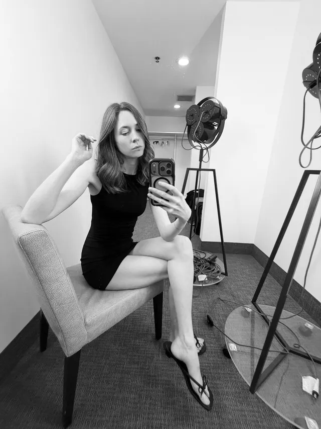 Karen Gillan sitting in a dark dress and heels for a mirror photo.