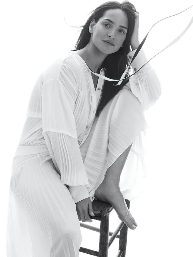 Adria Arjona sitting barefoot on a stool in a white outfit.