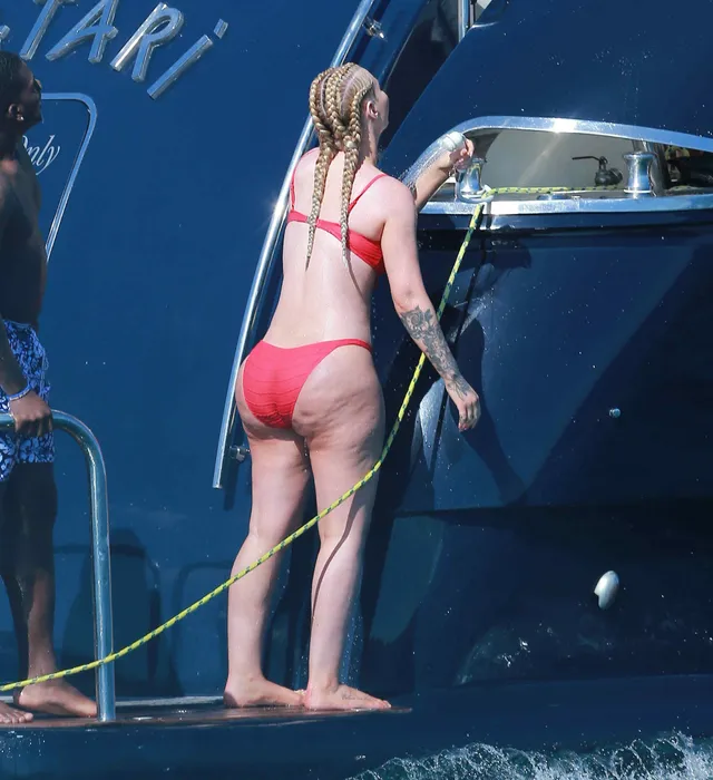 Iggy Azalea standing barefoot on a boat in a red swimsuit.