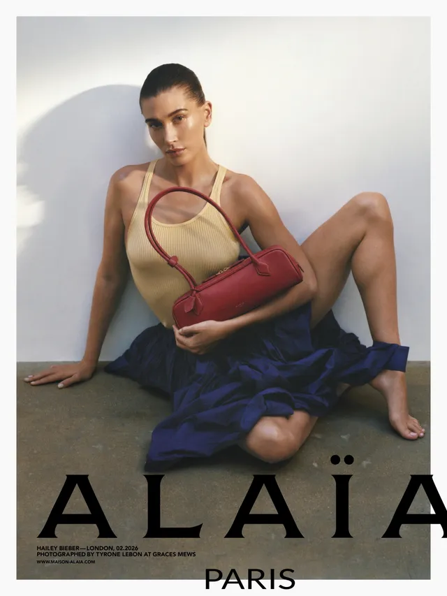 Hailey Bieber sitting barefoot in a studio holding a red handbag.