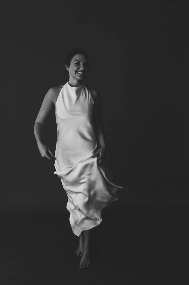 Gal Gadot posing barefoot in a flowing white dress against a dark background.