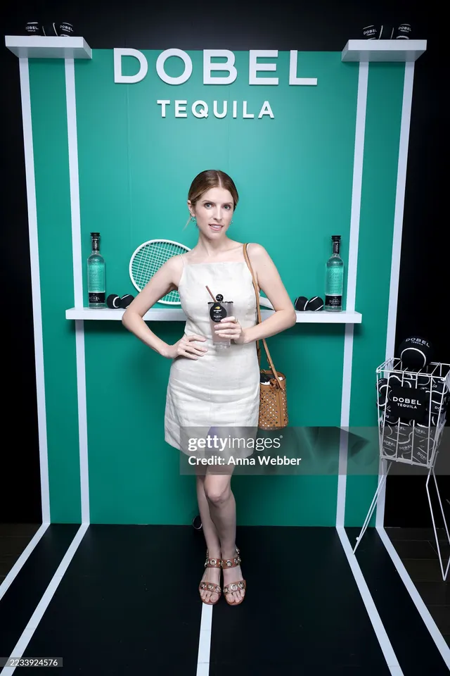 Anna Kendrick posing at an event in a silver dress and strappy heels.