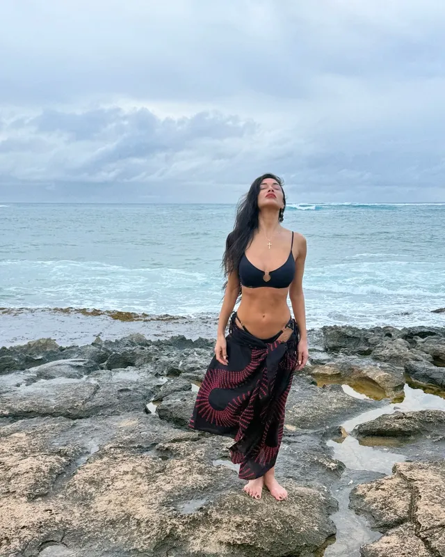 Nicole Scherzinger standing barefoot on coastal rocks beside the sea.