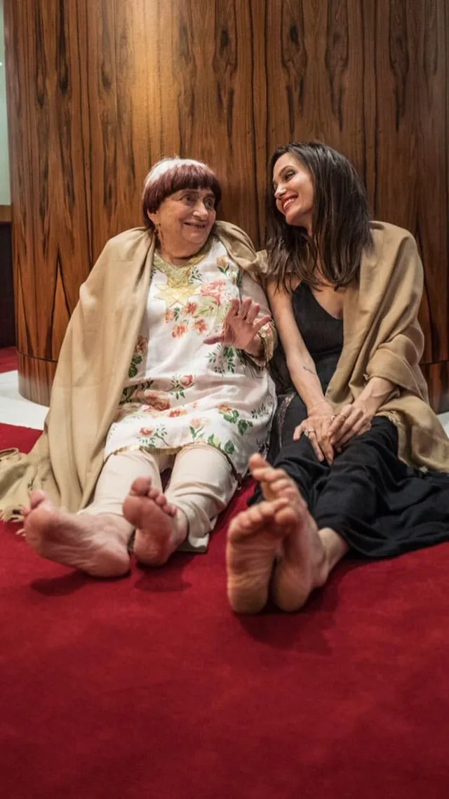 Angelina Jolie seated beside a friend on a red floor with bare soles visible.