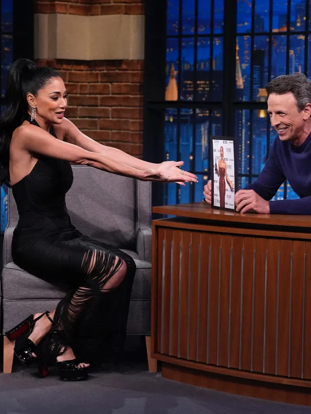 Nicole Scherzinger sitting during a television interview in a black dress and platform heels.