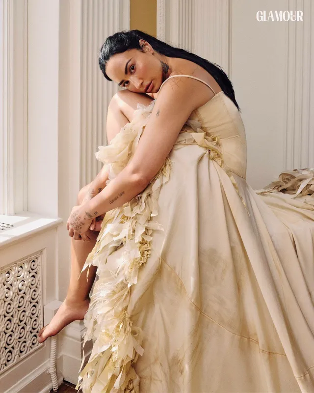 Demi Lovato sitting barefoot beside a window in a pale dress.