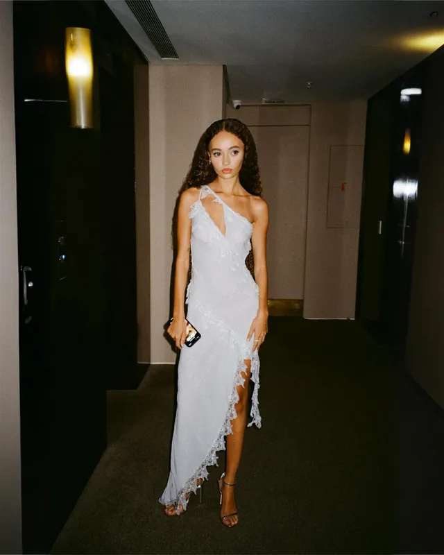 Nico Parker standing beside a mirror in a long pale dress and heels.