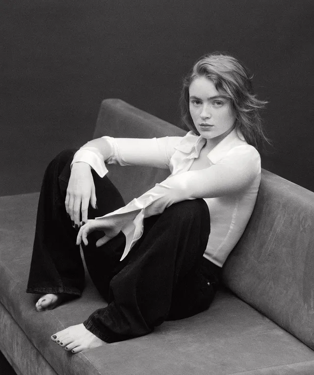 Sadie Sink sitting barefoot on a sofa in a monochrome portrait.