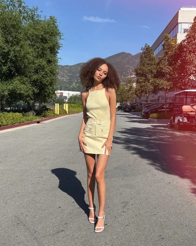 Nico Parker standing outdoors in a pale dress and white heels.