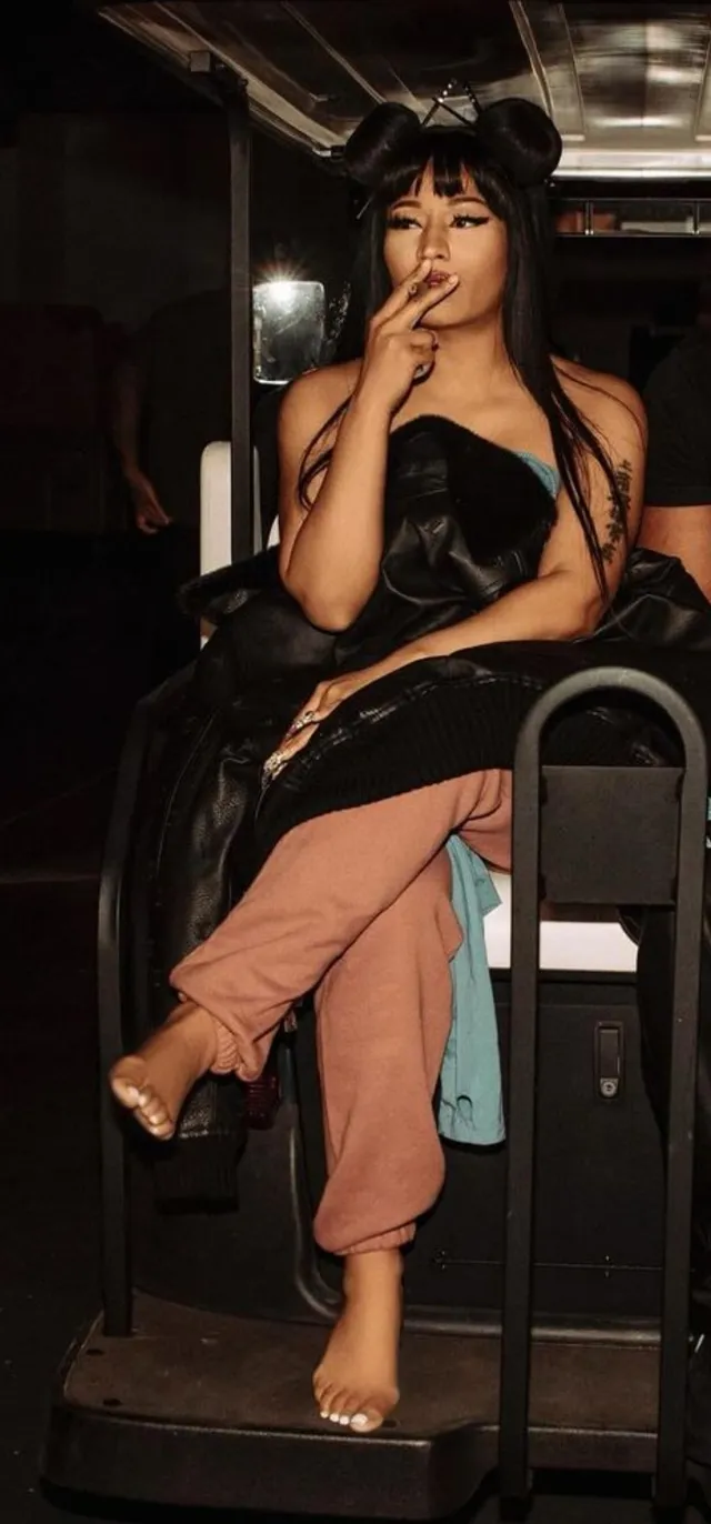 Nicki Minaj sitting in a director chair in a dark outfit and nude heels.