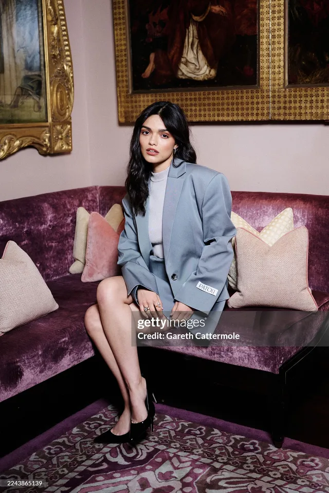 Rachel Zegler sitting on a purple sofa in a grey blazer dress and black heels.
