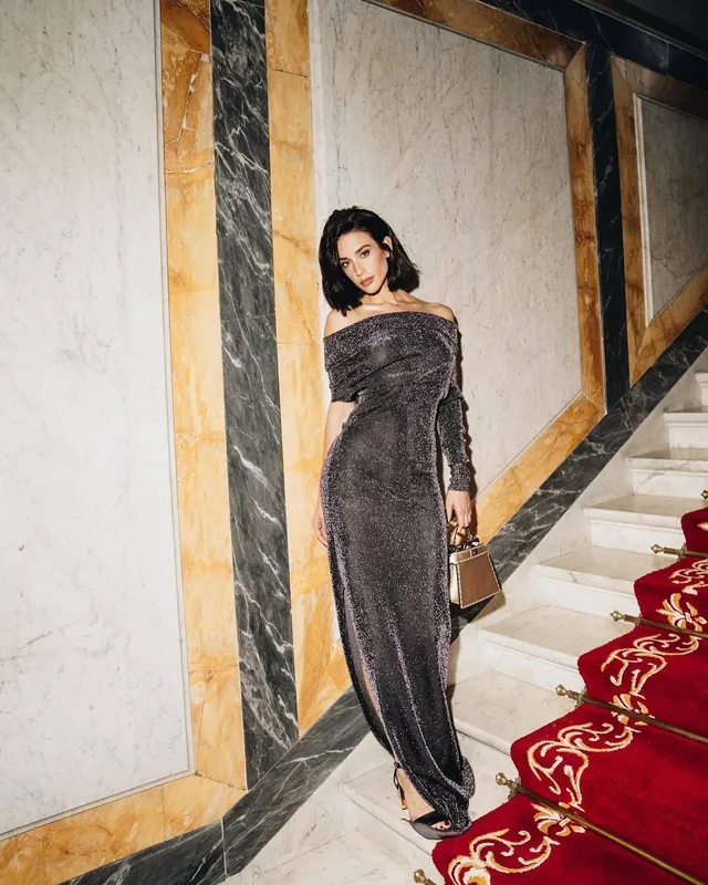María Pedraza wearing a long dark gown and black heels beside a staircase.