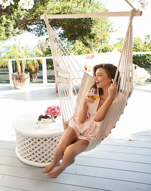 Miranda Kerr relaxing barefoot in a hanging chair on a porch.