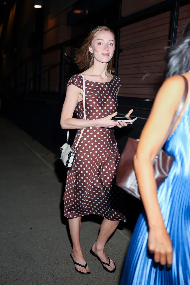 Phoebe Dynevor standing outdoors in a polka-dot dress and flip-flops.