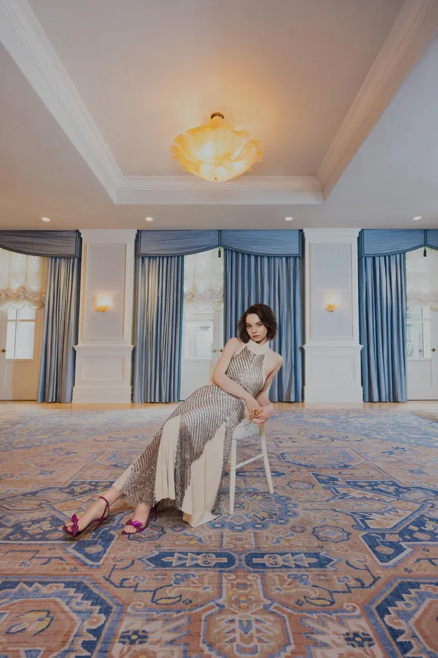 Emma Myers sitting in an armchair in a pale dress and red heels.