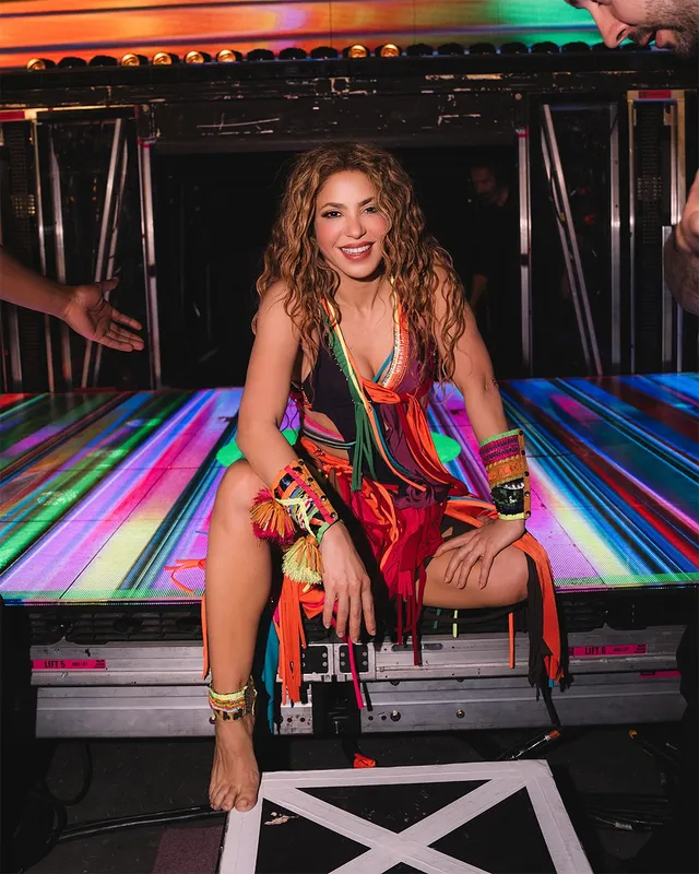Shakira sitting barefoot beside stage equipment wearing a colorful outfit.