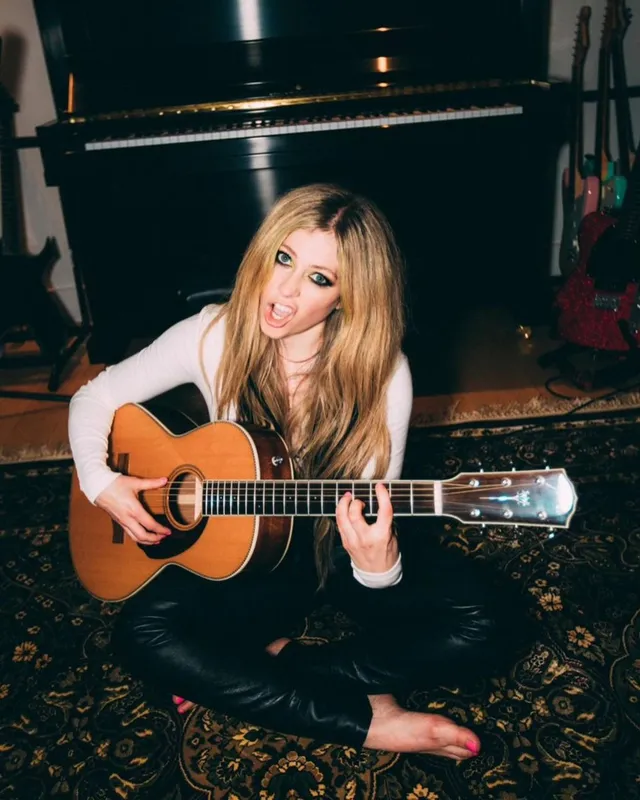 Avril Lavigne sitting barefoot on a rug while playing guitar.