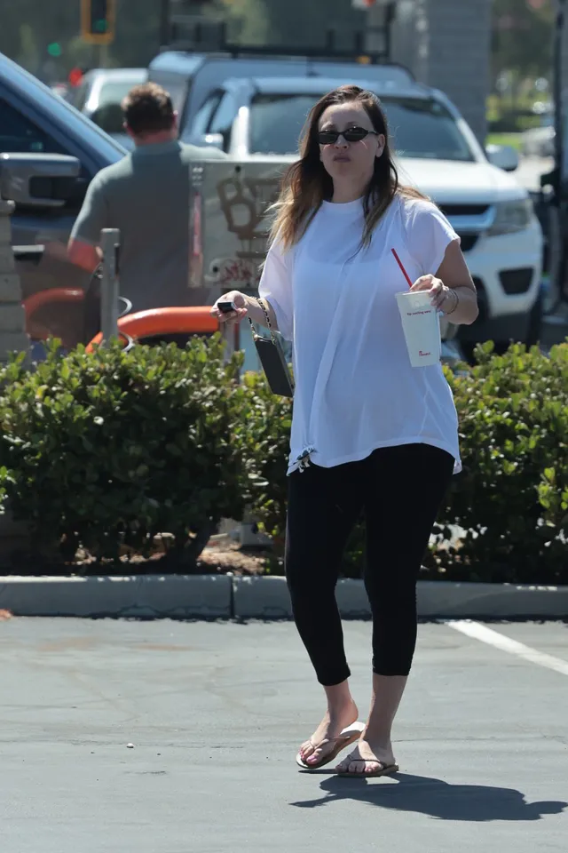 Kaley Cuoco walking outdoors in a white blouse, black leggings and flip-flops.