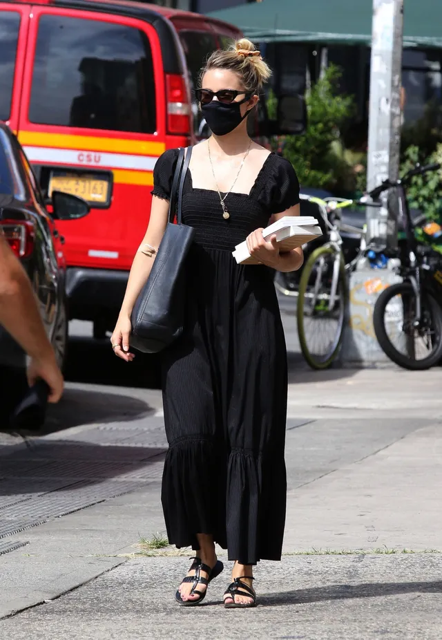 Dianna Agron walking outdoors in a black dress and flat sandals.