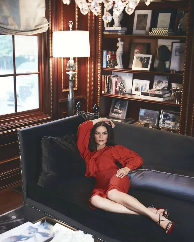 Catherine Zeta-Jones lounging on a dark sofa in a red outfit.