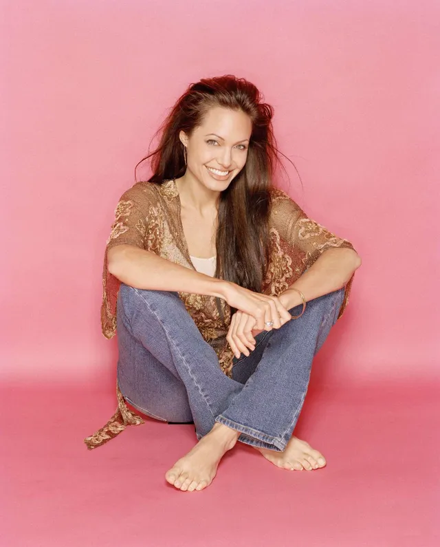 Angelina Jolie seated against a pink backdrop in jeans.