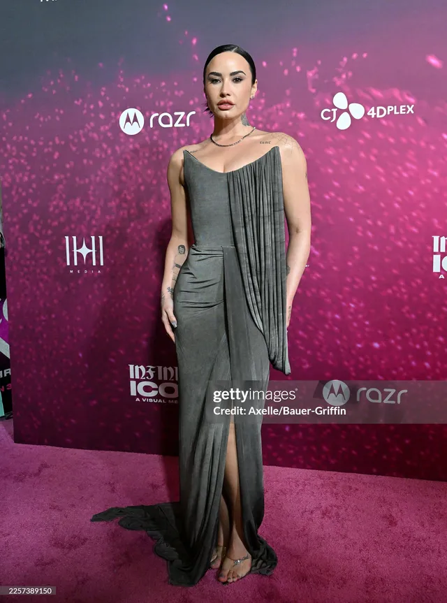 Demi Lovato standing at an event in a long grey dress and heels.
