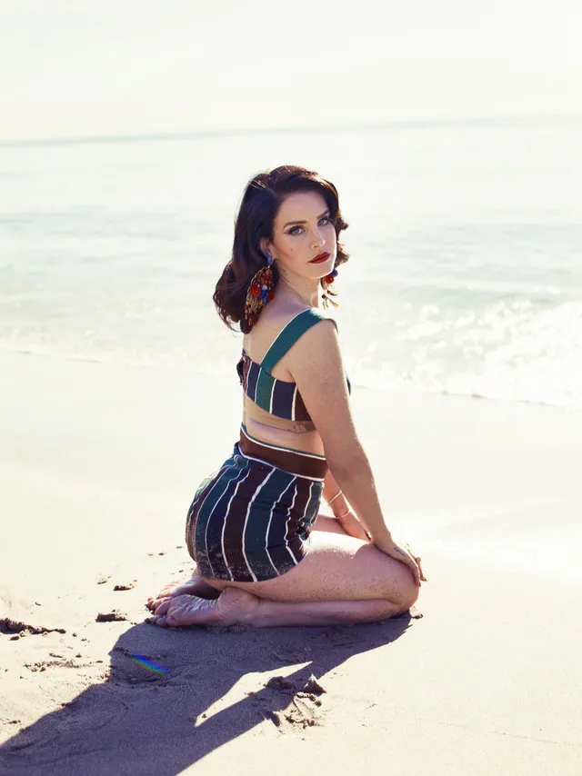 Lana Del Rey kneeling barefoot on a beach in a striped outfit.