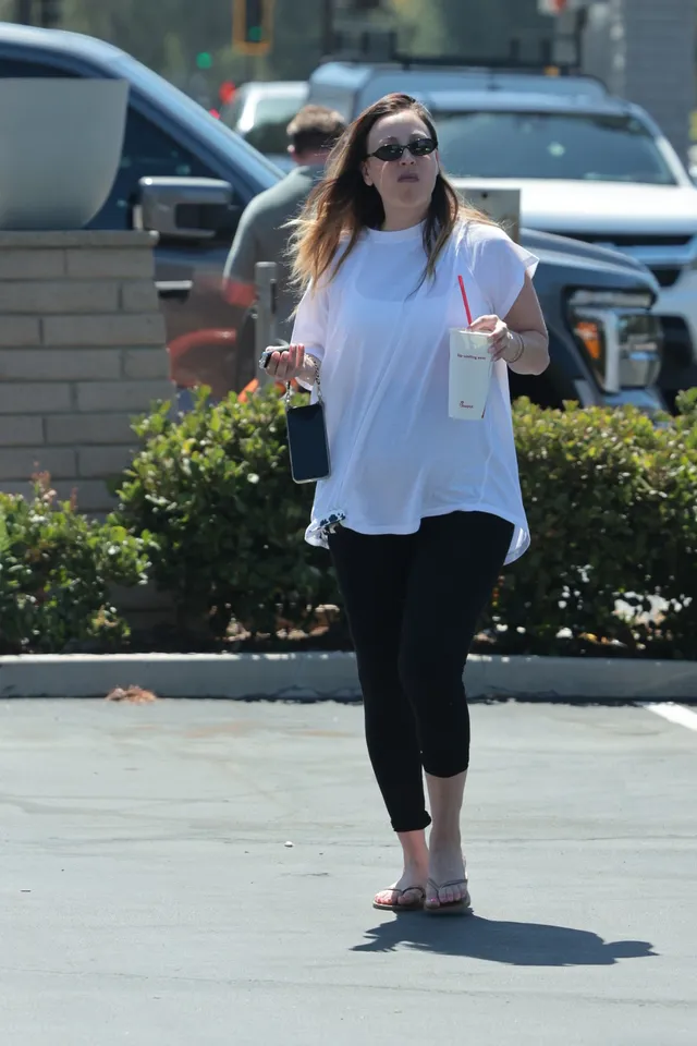 Kaley Cuoco standing outdoors in a white blouse and flip-flops.