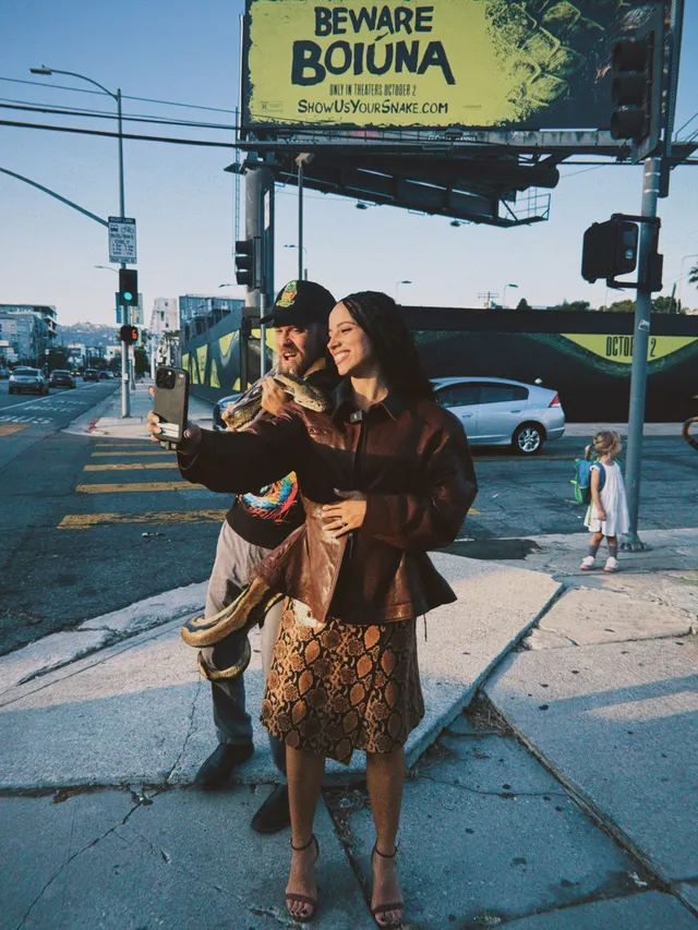Kiana Madeira posing with a companion on a sidewalk in a patterned dress and heels.