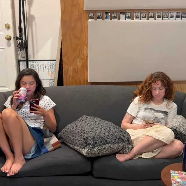 Olivia Rodrigo sitting barefoot on a sofa beside another woman.