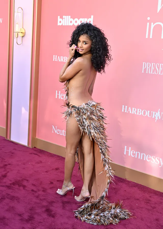 Tyla posing in clear heels and a feathered dress against a pink backdrop.