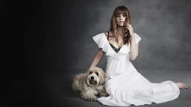 Alice Eve sitting in a white dress beside a dog in a studio portrait.