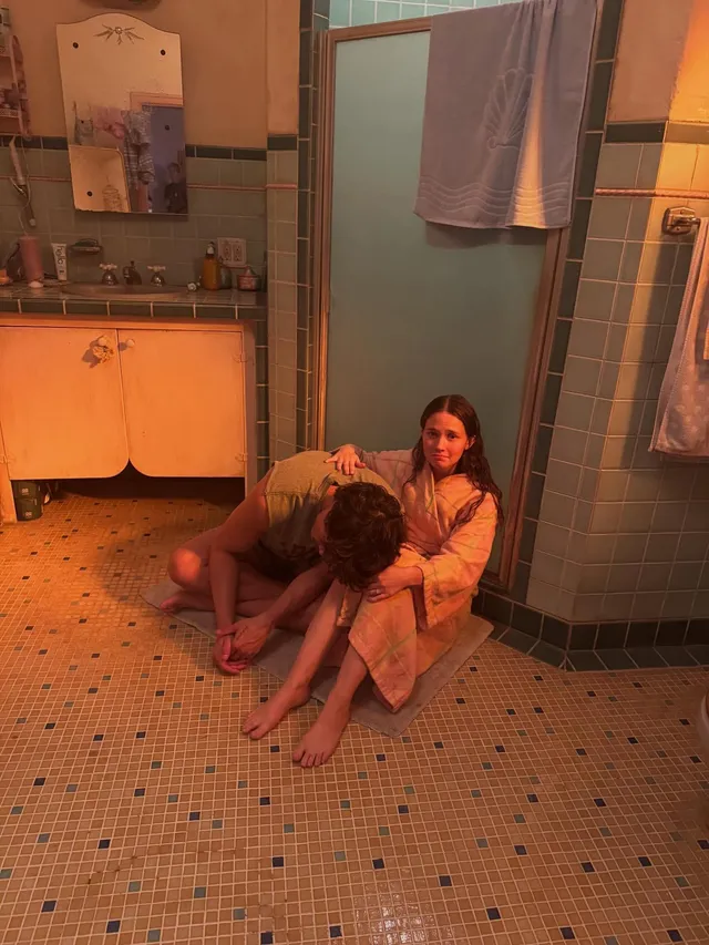 Cailee Spaeny sitting barefoot beside another person on a bathroom floor.