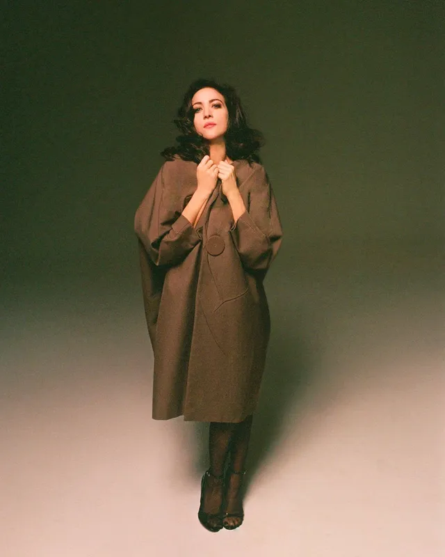 Brittany Snow posing for a studio portrait in a long coat and boots.