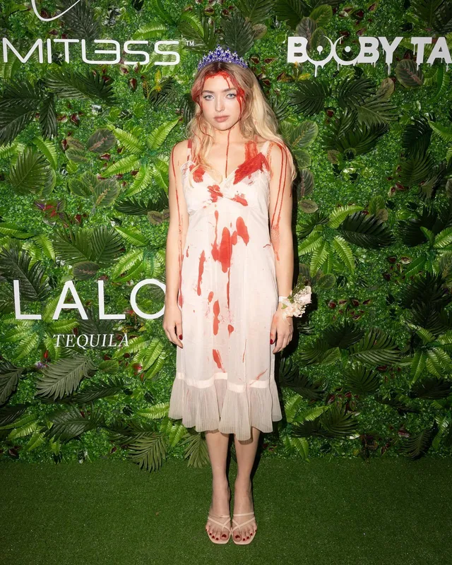 Peyton List standing in a pale dress and heels beside a leafy event backdrop.