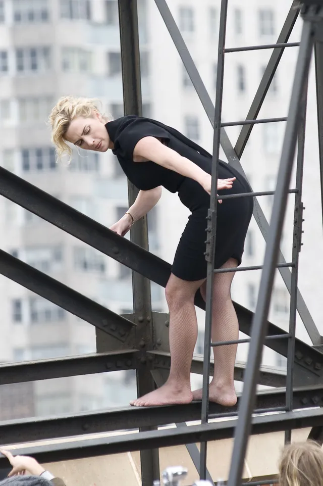 Kate Winslet standing barefoot on an outdoor staircase in a black dress.