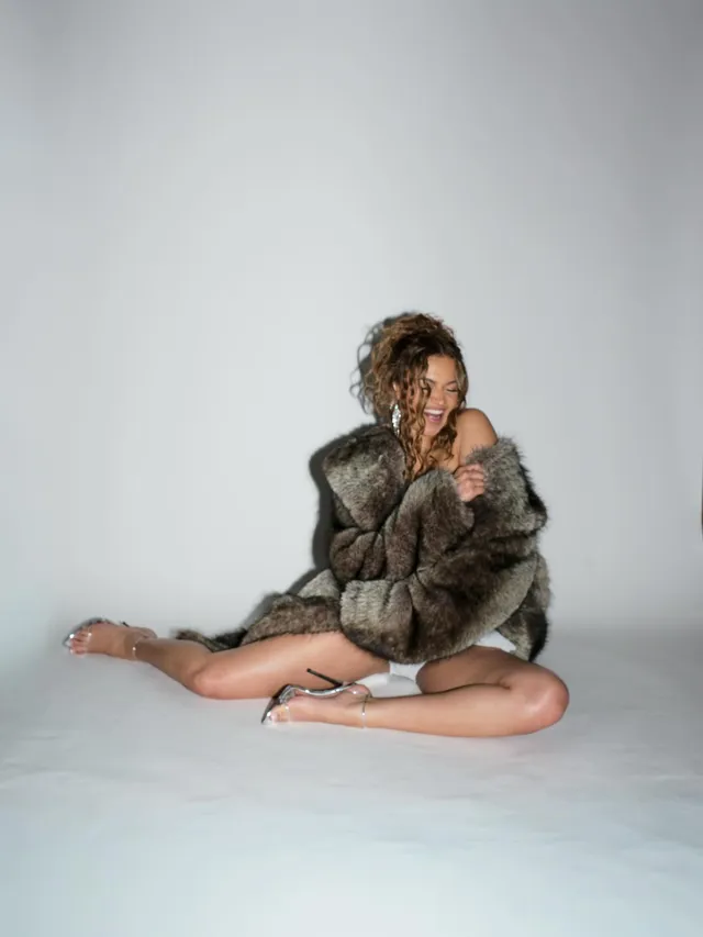 Madison Bailey sitting on a studio floor in a fur coat and clear heels.